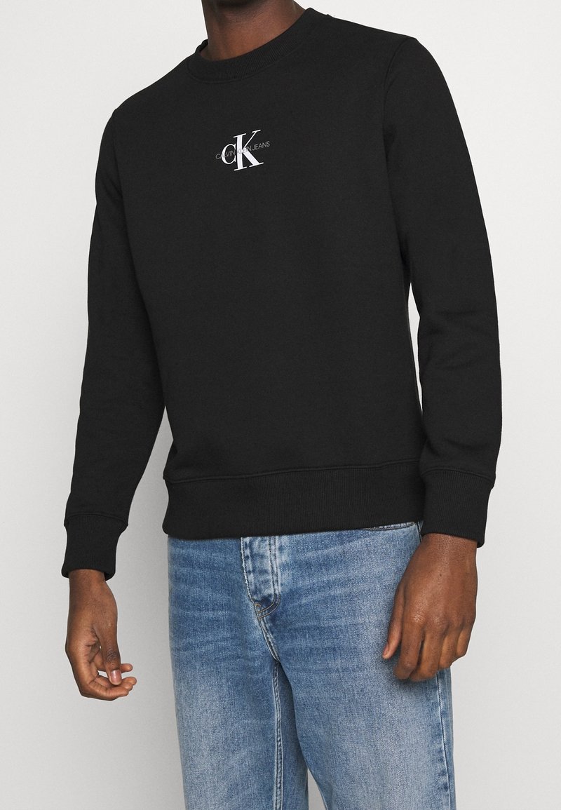 Homme portant un sweat-shirt noir Calvin Klein avec un logo blanc et un jean bleu clair, se tenant devant un fond uni.