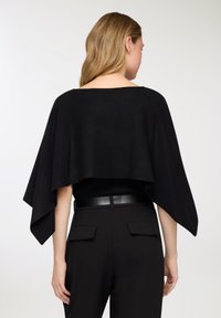 Poncho nero in maglia con design ampio e fluente. Presenta una scollatura a barca e cade sulle spalle, abbinato a pantaloni neri a vita alta.