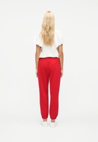 Γυναίκα με μακριά ξανθά μαλλιά στέκεται γυρισμένη πλάτη, φοράει λευκό μπλουζάκι με κοντά μανίκια, κόκκινο παντελόνι τζόγκινγκ και λευκά αθλητικά παπούτσια σε απλό φόντο.