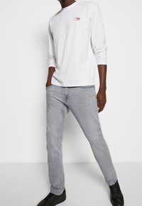 Camiseta de algodón blanca de manga larga con el logo rojo "TOMMY JEANS", combinada con jeans de mezclilla gris claro y zapatillas negras. Diseño simple y moderno.