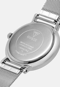 Reloj de acero inoxidable con correa de malla. La parte trasera presenta el logo "GUESS", detalle de resistencia al agua y número de modelo grabado. Superficie pulida.