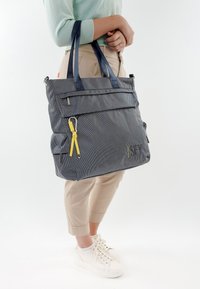 Persona con pantaloni beige e sneakers bianche che tiene una grande borsa tote grigia con cinturini blu navy e una cerniera gialla.