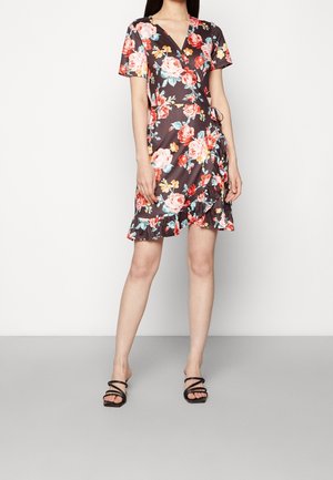Femme portant une robe portefeuille noire à fleurs avec manches courtes et ourlet volanté, associée à des sandales à talons noires à brides sur un sol gris.