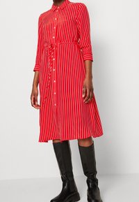 Vestito a righe rosse e bianche con bottoni, vita con lacci, maniche lunghe e orlo al ginocchio, abbinato a stivali neri al ginocchio.