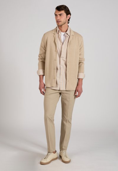 Homme debout portant un blazer beige, une chemise rayée beige et blanche, un pantalon beige et des baskets crème sur un fond clair uni.