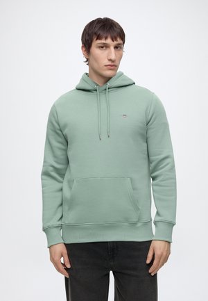 Jeune homme portant un sweat à capuche vert clair avec une poche à l'avant et un pantalon noir, debout devant un fond blanc uni.