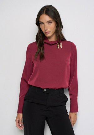 Mujer con cabello largo y oscuro vistiendo una blusa burdeos de manga larga con detalles decorativos dorados y pantalones negros de tiro alto sobre un fondo liso.