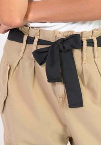Beige broek van textuurstof, met een brede zwarte stoffen strikriem. De tailleband heeft riemlussen en zijzakken.