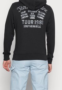Hombre vistiendo una sudadera negra con texto blanco que enumera los estados de EE.UU. y "Tour 1981 Southern Ride" en la parte posterior, combinada con pantalones cargo de color azul claro.