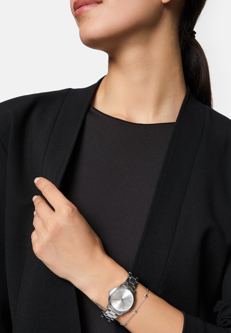 Silberne Armbanduhr mit rundem Ziffernblatt, metallisch-glänzendem Armband und minimalistischer Gestaltung. Am Handgelenk über einer schwarzen Jacke und einem Oberteil getragen.