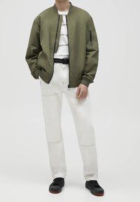 Olijfgroene bomberjack met een ritssluiting aan de zak, gedragen over een wit t-shirt met strepen. Gecombineerd met witte broek met patchdetails en zwarte sneakers.