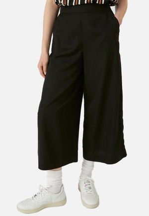 Osoba ubrana w szerokie, czarne spodnie o długości 7/8, białe skarpetki i białe trampki, stojąca z jedną ręką w kieszeni.