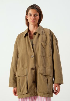 Frau trägt eine lockere, geknöpfte braune Jacke mit großen Fronttaschen über einem gestreiften rosa-weißen Rock oder Hose vor einem einfarbigen Hintergrund.