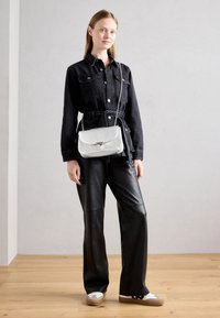 Giacca di jeans nera con due tasche e cinturino in vita, abbinata a pantaloni in pelle nera. Il modello porta una borsa a tracolla metallica argentea.