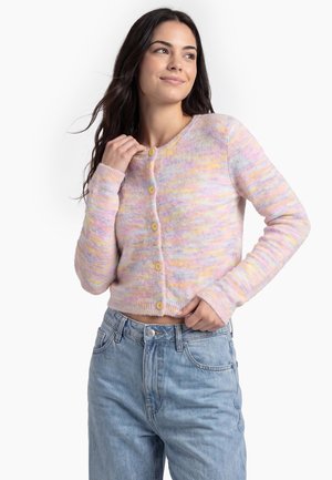 Ung kvinde iført en pastel flerfarvet knappet cardigan og lyseblå jeans, poserer med den ene hånd ved nakken og den anden på taljen.
