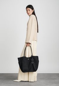 Μαύρη τσάντα tote από υφασμά με υφή νάιλον, με διπλούς χειρολαβούς, εξωτερικές τσέπες και ένα γούρι. Το μοντέλο φοράει ένα υπερμεγέθες μπεζ πουλόβερ και κρεμ παντελόνι.