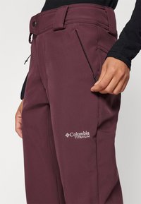 Columbia Titanium tummanpunaiset housut sileällä pinnalla, jossa on sivupokkari ja logo reidessä, kestävä kangas, joka on suunniteltu kestäväksi.