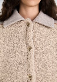 Giacca di pile beige con colletto in feltro beige. Presenta grandi fibre testurizzate e quattro grandi bottoni rotondi sul davanti.
