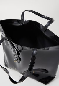 Svart lackläder toteväska med två handtag, öppen topp och guldlogotypdetalj. Slät, glänsande yta och strukturerad form.