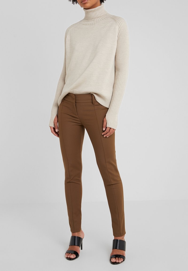 Pull col roulé beige avec des manches côtelées, associé à un pantalon marron ajusté. La personne se tient debout dans des sandales à talons carrés noires.
