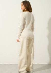 Jasno beżowy sweter o dopasowanym kroju i długich rękawach, wykonany z miękkiej tkaniny. W połączeniu z luźnymi, kremowymi spodniami.