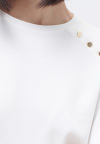 Top de algodón blanco con cuello redondo y botones a presión en tono dorado en el hombro. Textura suave con un diseño limpio y minimalista.