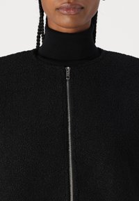 Black fleece jacket with a high collar, featuring a front zipper and textured surface. Simple, unembellished design.