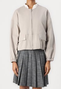 Chaqueta bomber recortada de color beige claro con cremallera, puños elásticos y bolsillos frontales, combinada con una falda gris con pliegues. Textura de tela suave.