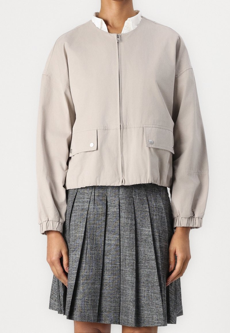 Chaqueta bomber recortada de color beige claro con cremallera, puños elásticos y bolsillos frontales, combinada con una falda gris con pliegues. Textura de tela suave.