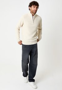 Pull en tricot beige clair avec un col montant et une fermeture éclair partielle, associé à un jean droit gris foncé et des baskets blanches.