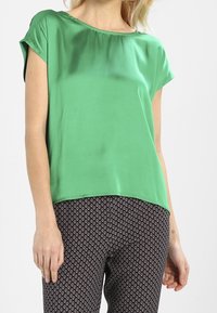 Top de satén verde con escote barco y mangas cortas, combinado con pantalones de patrón en blanco y negro. Textura suave, corte relajado.