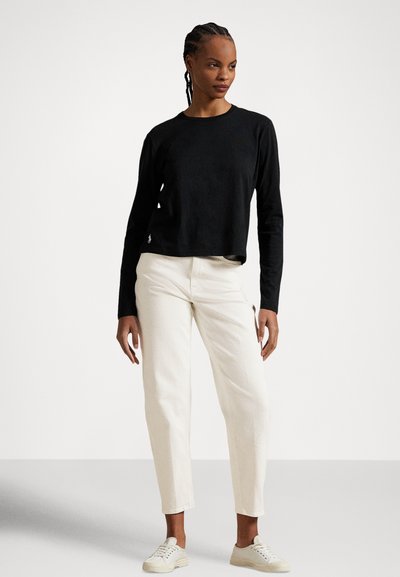 Frau mit geflochtenen Haaren, die ein schwarzes langärmeliges Top, weiße Hose mit hoher Taille und weiße Sneakers trägt und vor einem einfarbigen Hintergrund steht.