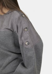Donna che indossa un maglione grigio a maniche lunghe con un pannello grigio più chiaro sulla spalla, caratterizzato da tre grandi bottoni.