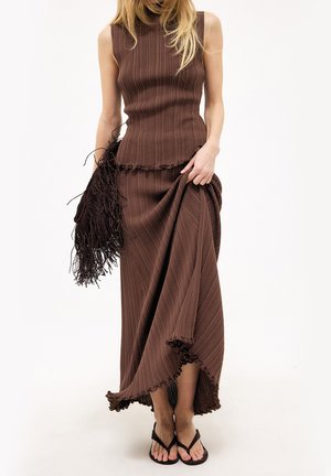 Femme portant un haut marron sans manches en maille côtelée et une jupe longue assortie, tenant la jupe et un sac marron à franges, avec des sandales noires.