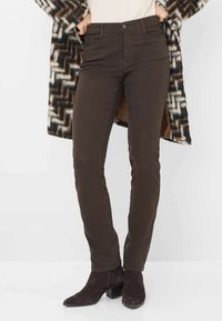 Jeans slim-fit marroni con tasche, chiusura a bottone e dettagli di cucitura sottili, abbinati a stivaletti neri e un cardigan a fantasia.