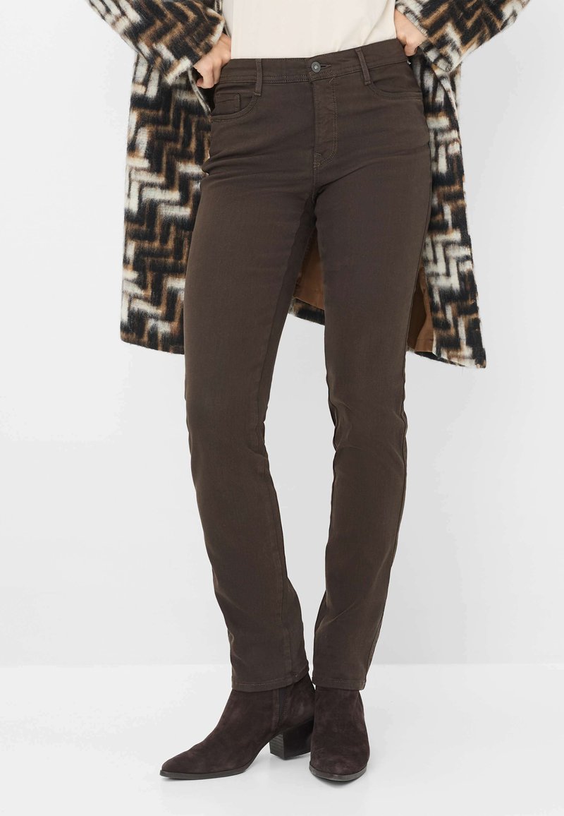 Jeans slim-fit marroni con tasche, chiusura a bottone e dettagli di cucitura sottili, abbinati a stivaletti neri e un cardigan a fantasia.