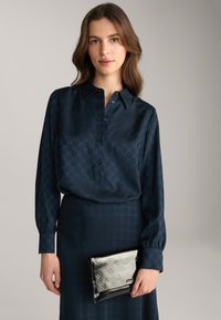 Blusa di seta blu navy con un sottile motivo geometrico, chiusura frontale con bottoni e maniche lunghe, abbinata a una borsa a tracolla nera lucida con design in rilievo.