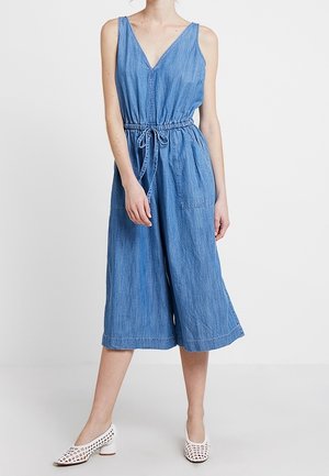 Combinaison sans manches en denim bleu avec décolleté en V et taille à cordon, portée avec des chaussures blanches perforées à petit talon sur une personne debout.