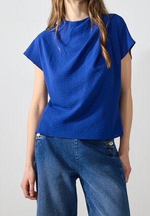Kobieta w teksturowanym, królewskim niebieskim topie z krótkim rękawem, w połączeniu z wysokim stanem i szerokimi nogawkami niebieskich dżinsów, na jednolitym tle.