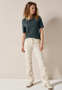 Mörkgrön t-shirt med kort ärm, kombinerad med krämfärgade utsvängda jeans och ljusa sneakers, mot en neutral bakgrund.