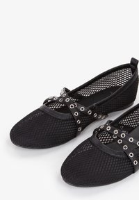 Bailarinas negras de malla con punta redondeada, con correas entrelazadas adornadas con ojales plateados. Superior texturizado y suela negra sólida.