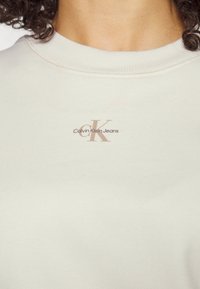 Felpa girocollo color crema realizzata in tessuto morbido, con un logo marrone con la scritta "Calvin Klein Jeans" stampata sul davanti.