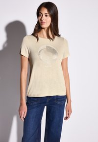 T-shirt beige manica corta con una grafica circolare argentata che riporta la scritta "SOLEIL" al centro. Abbinalo a jeans di denim blu scuro. Texture liscia.