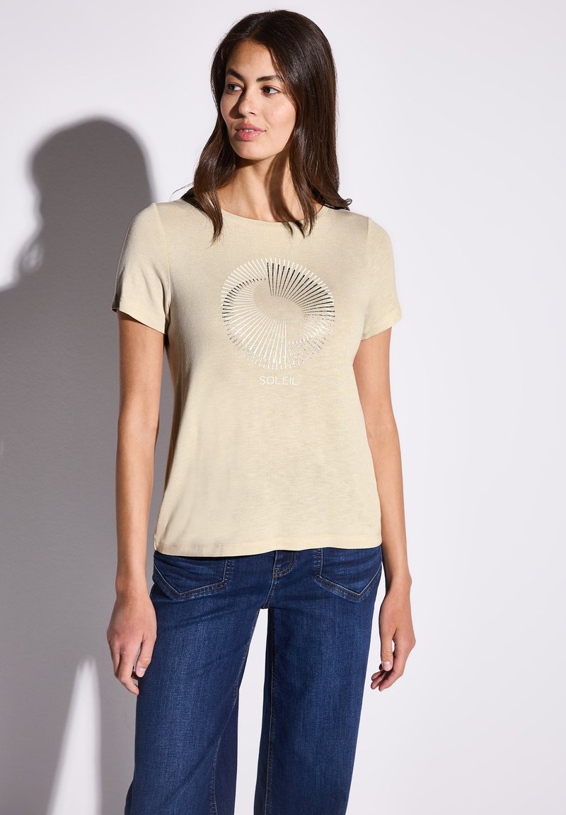 T-shirt beige manica corta con una grafica circolare argentata che riporta la scritta "SOLEIL" al centro. Abbinalo a jeans di denim blu scuro. Texture liscia.