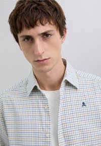 Koszula w kratę z lekkiego materiału, z wzorem w niebiesko-brązową kratkę. Zapinana na guziki, z małym granatowym logo w kształcie czaszki na piersi.