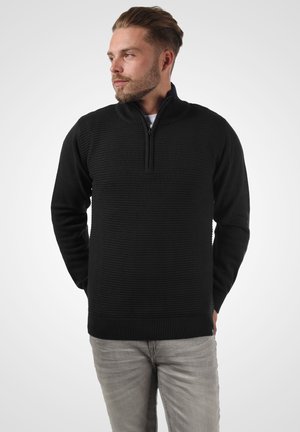 Black knitted pullover with a zip neck, featuring a textured design. Long sleeves and ribbed cuffs and hem. Worn with light gray pants.