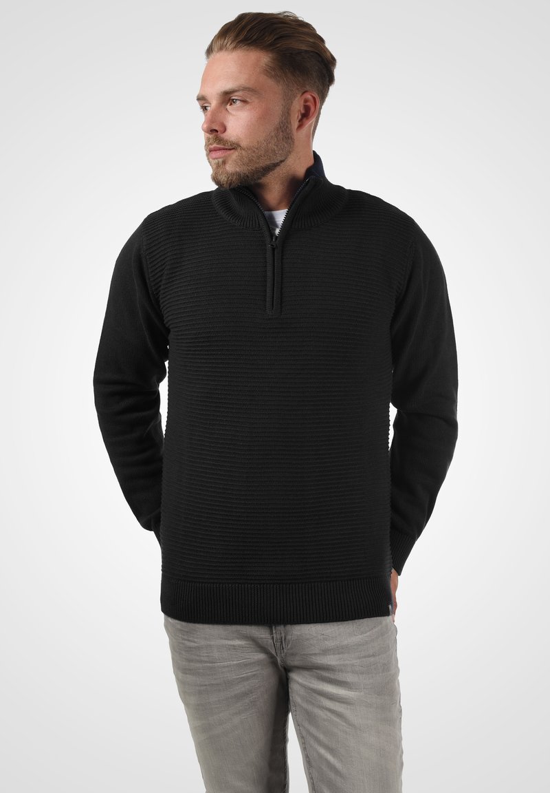 Schwarzer gestrickter Pullover mit Reißverschlusskragen, der ein strukturiertes Design aufweist. Lange Ärmel und gerippte Bündchen sowie Saum. Getragen mit hellgrauen Hosen.