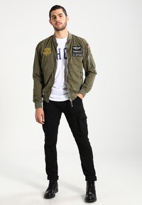 Olijfgroene bomberjack met patches, voorrits, zijzakken en geribbelde manchetten. Draagt over een wit shirt en gecombineerd met zwarte cargobroek.