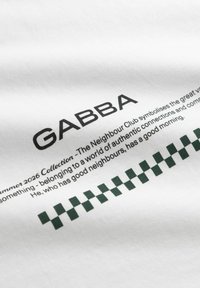 Tissu blanc avec le texte "GABBA", une petite description imprimée, et un motif à damier vert foncé près du bord inférieur.