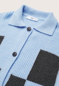 Světle modrý strukturovaný kardigan s límcem, který má černé obdélníkové nášivky a dva černé knoflíky vpředu. Viditelná značka.